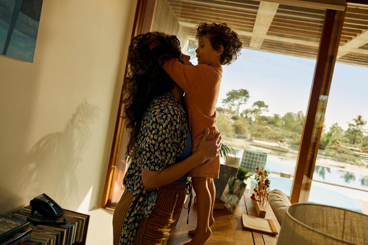 A woman stands in a living room supporting a toddler who is standing on a table. The toddler is playing with her hair.